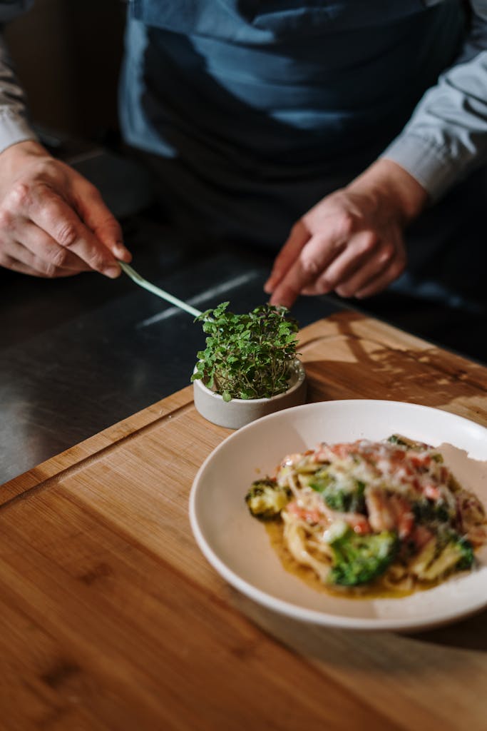 Close-up of a chef adding fresh herbs to a plate of gourmet pasta in a kitchen setting.